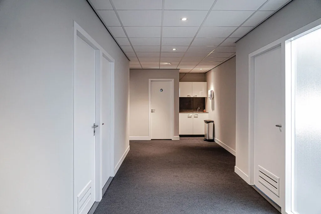 Witte, strak afgewerkte gang met meerdere deuren en een kitchenette aan het einde, in een pand aan de Burgemeester van Karnebeeklaan.